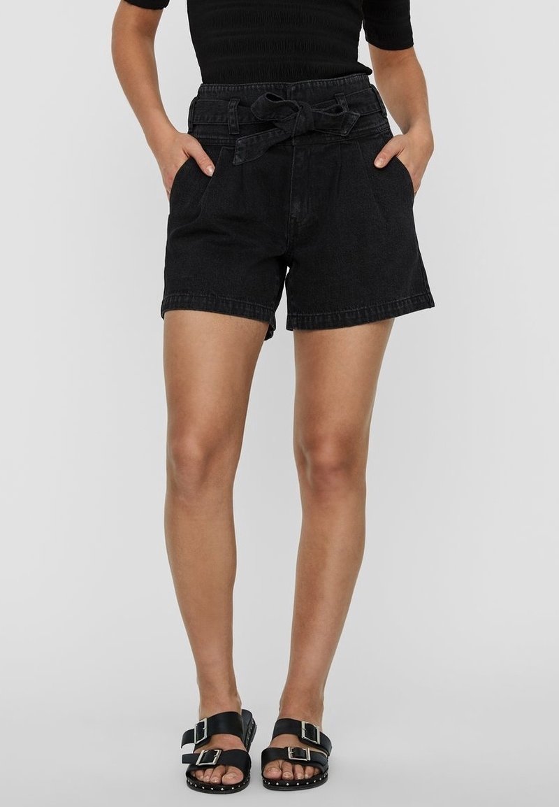 Femme portant un short noir ceinturé, un top noir et des sandales noires à deux boucles, debout les mains dans les poches.