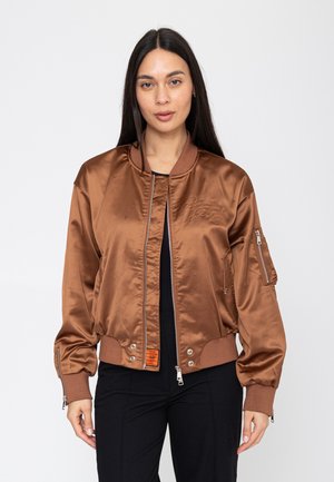 Vrouw met lang donker haar die een koperen satijnen bomberjack draagt over een zwarte broek, staand tegen een effen witte achtergrond.
