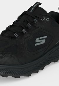 Μαύρο αθλητικό παπούτσι Skechers με κορδόνια, υφασμάτινη, αδιάβροχη σόλα και ανακλαστικό λογότυπο "S" στο πλαϊνό πάνελ.