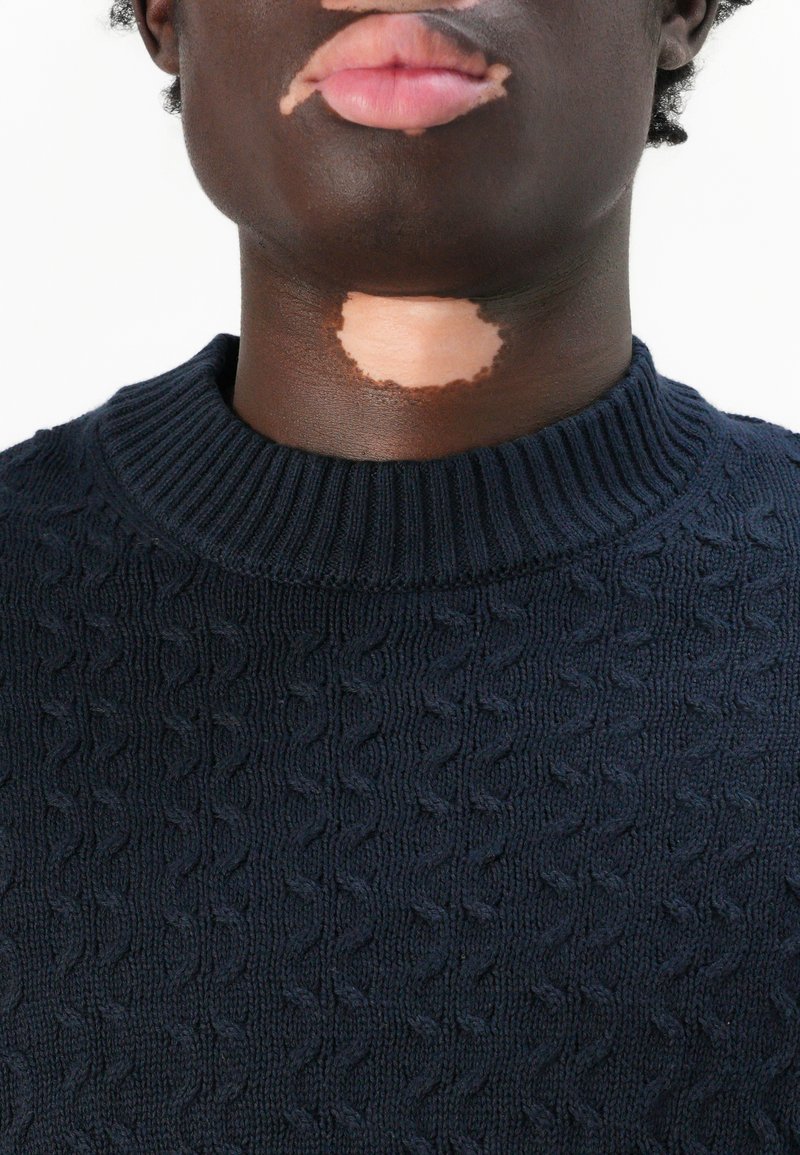 Pull en maille bleu marine avec un col côtelé, présentant un motif texturé. La peau montre une tache plus claire sous le menton, se fondant dans un teint de peau plus foncé.