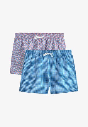 Giovane uomo in piedi contro un muro bianco texture, con una maglietta bianca senza maniche, pantaloncini blu con fantasia e sandali marroni.
