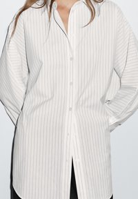 Camicia bianca a maniche lunghe con strisce verticali blu navy, chiusura con bottoni, vestibilità ampia e spacchi laterali, realizzata in tessuto leggero.