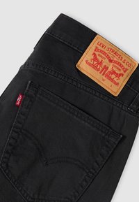 Μαύρο παντελόνι από τζιν με καφέ δερμάτινο πίσω ταμπελάκι που φέρει το λογότυπο Levi Strauss. Περιλαμβάνει κόκκινη ετικέτα και χαρακτηριστική ραφή στην τσέπη.