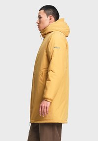Homme portant un manteau d'hiver à capuche jaune moutarde avec le logo "AIGLE" sur la manche, debout de profil sur un fond uni.