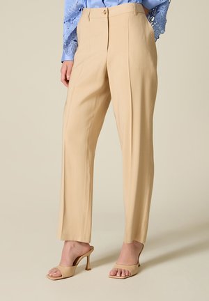 Beige skræddersyede bukser med lige ben kombineret med matchende hælede sandaler og en blå top med blonder ærmer, vist fra taljen til fødderne.