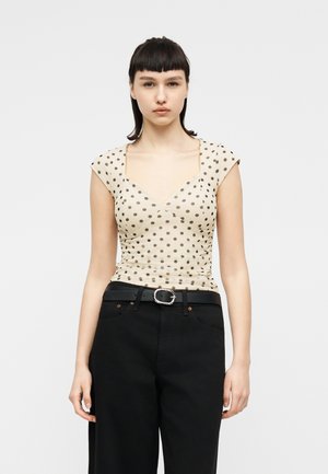 Ung kvinne med kort, mørkt hår iført en beige polkaprikket topp med hette og sorte høytlivsbukser med belte.