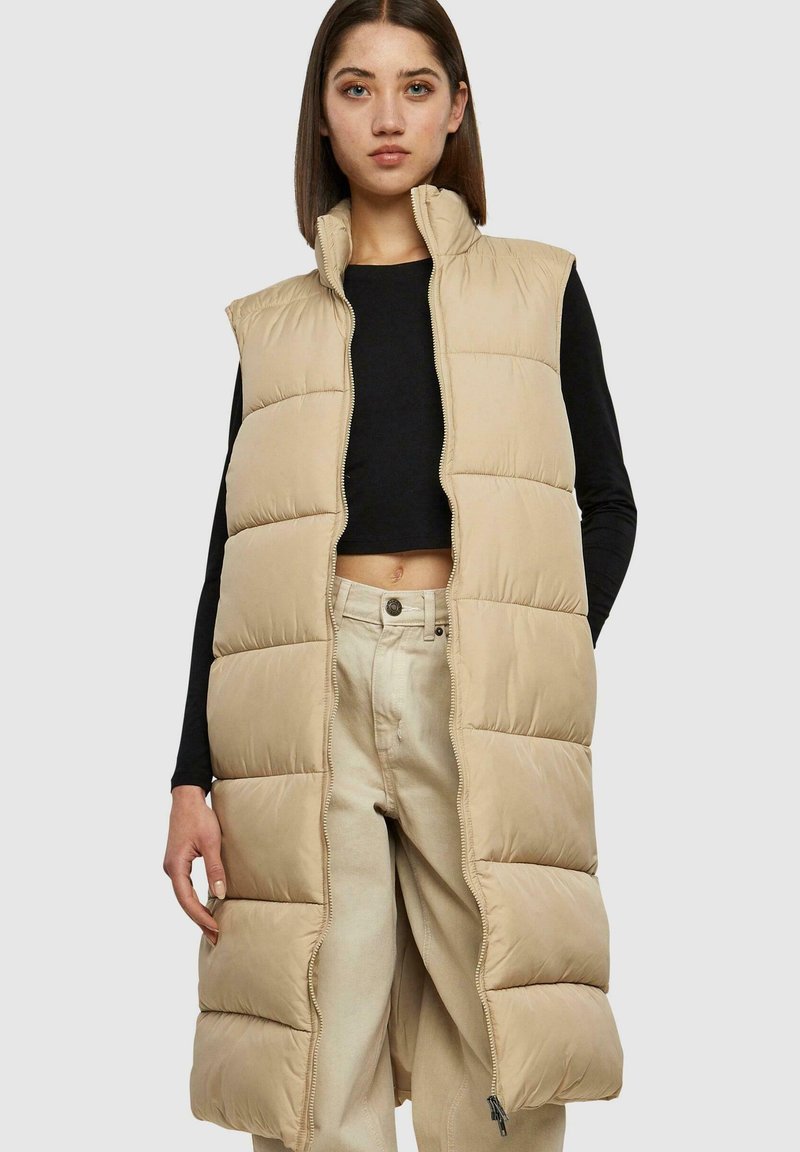 Beige gewatteerde vest met hoge kraag, ritssluiting en zonder mouwen, gedragen over een zwarte top, met een gladde textuur en een doorgestikt patroon.