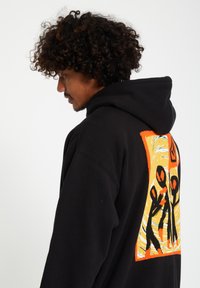 Zwarte hoodie met een kleurrijk, abstract ontwerp op de achterkant, featuring stickfiguren en spiralen, gemaakt van zachte stof, met een ontspannen pasvorm.