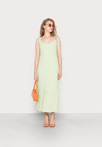 Vestido midi verde com alças com rendas, tecido texturizado e parte inferior rodada; combinado com uma bolsa laranja e sandálias. Óculos de sol redondos usados.