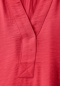 Röd topp med V-ringning, tillverkad av ett texturerat tyg. Har en enkel design med subtila sömmar och en tab-detalj nära ringningen.
