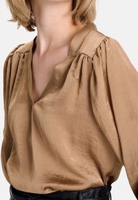 Blouse en satin marron avec un col en V profond, un détail plissé aux épaules et une texture douce et lisse. Présente un drapé élégant et une coupe décontractée.