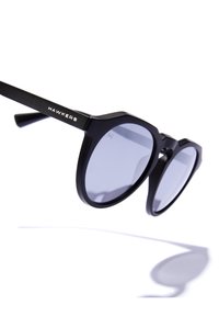 Mustat aurinkolasit, joissa on pyöreät linssit, mattapinta ja rohkea geometrinen muoto. Brändin nimi "HAWKERS" on kaiverrettu temppeliin.