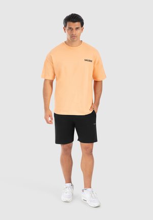 Lys orange t-shirt med sort logo "SMILODOX," korte ærmer, parret med sorte shorts og hvide sneakers, stående på en neutral baggrund.