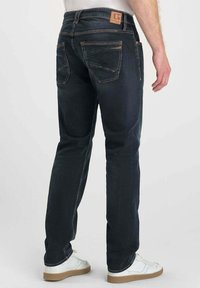 Jean en denim bleu foncé avec une coupe droite, poches arrière visibles et patch en cuir marron à la taille. Portés avec des baskets blanches.