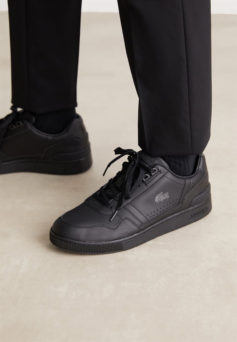 Black leather sneakers with a textured surface, featuring round laces, perforated accents, and the Lacoste logo on the side.