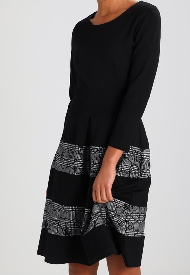 Robe noire à manches longues, avec un corsage ajusté et une jupe évasée. La jupe comporte des bandes argentées à motifs avec des détails en dentelle et en carreaux.