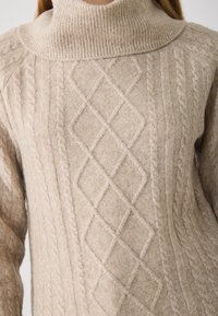 Pull en maille torsadée beige à col roulé avec des motifs texturés, y compris des designs en tresses et en losanges. Matière douce et confortable ressemblant à de la laine.