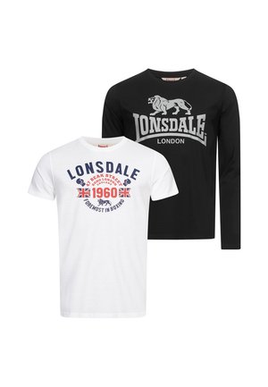 Młody mężczyzna w białej koszulce Lonsdale z grafiką, czarnych dżinsach, białych trampkach, trzymający granatową czapkę Lonsdale w prawej ręce.