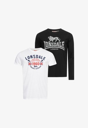 Jeune homme portant un t-shirt blanc à motif Lonsdale, jean noir, baskets blanches, tenant une casquette Lonsdale bleu marine dans la main droite.