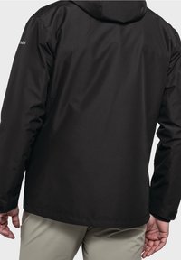 Černá kapucí nošená osobou s béžovými kalhotami, viditelné logo značky na levém rukávu, ruce uvolněně po stranách na jednobarevném pozadí.