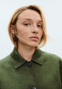 Cappotto in suede verde con colletto e due bottoni neri; il modello ha i capelli biondi corti e grandi orecchini rotondi. Trucco naturale e sfondo neutro.