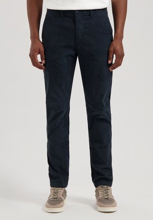 Dunkelblaue Baumwollchinos mit geradem Bein, frontalen Taschen und einer glatten Textur. Kombiniert mit hellgrauen Wildledersneakern.
