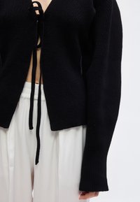 Μαύρο πλεκτό ζακέτα δεμένο χαλαρά μπροστά πάνω από γυμνή κοιλιά, συνδυασμένο με λευκό φαρδύ παντελόνι σε απλό φωτεινό φόντο.