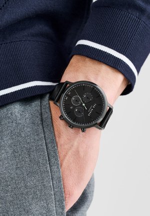 Black analog watch with a textured leather strap, round black dial, three sub-dials, and silver accents. Worn on a wrist with gray fabric.