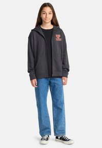 Grijze zip-up hoodie met oversized pasvorm, voorzakken, en "RIDE THE RAILS" in het rood gedrukt. Gecombineerd met blauwe rechte spijkerbroek en zwarte sneakers.
