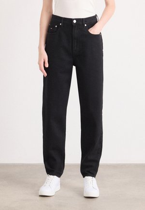 Personne portant un jean noir taille haute à jambes droites avec un bouton blanc et des baskets blanches, debout sur un sol gris clair.