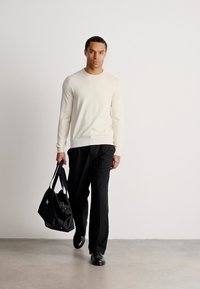 Mann, der geht, trägt einen cremefarbenen Pullover, schwarze Hose und schwarze Schuhe und hält eine schwarze Tragetasche in der linken Hand vor einem schlichten weißen Hintergrund.
