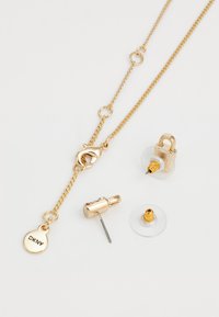Gouden sieradenset bestaande uit een kettinghalsketting met een sluiting en een label, plus oorbellen met gemengde details en transparante achterkant.