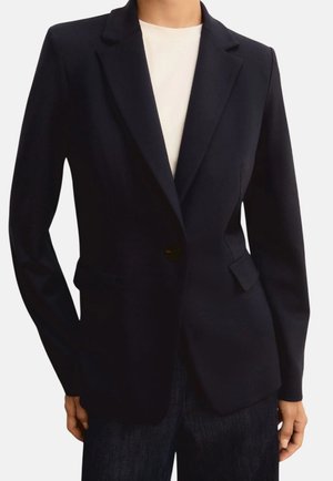 Donna che indossa un blazer nero aderente con un solo bottone, rever a incavo e tasche a pattina sopra una canottiera bianca e pantaloni scuri.