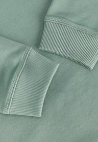 Gros plan des manches d'un sweat-shirt vert montrant des poignets côtelés et des coutures détaillées sur un tissu doux.