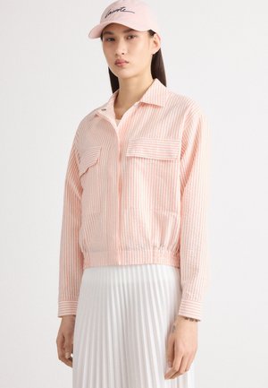 Gestreepte, lichtgewicht, roze jas met twee voorzakken en een gerimpelde zoom. Gecombineerd met een roze pet met zwart geborduurde tekst.