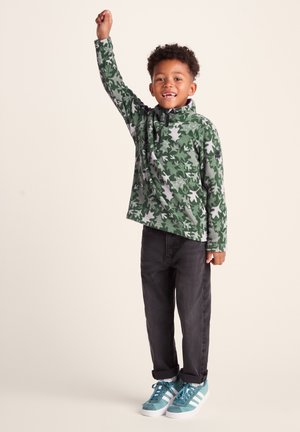 Green fleece pullover with white leaf pattern, half-zip collar; paired with dark jeans and teal sneakers with white accents.