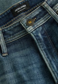 Közeli kép sötétkék farmernadrágról, amelyen gomb, övbújtató és fekete JACK & JONES címke látható fehér felirattal a derékrészen.