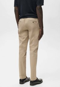 Beige chino's met een rechte snit, voorzien van achterzakken en subtiele stikdetails. Gecombineerd met zwarte schoenen en een donker shirt.