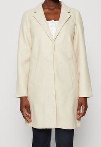Donna che indossa un cappotto lungo color crema con revers, sopra una camicia bianca con bottoni e pantaloni scuri, in piedi su uno sfondo neutro.