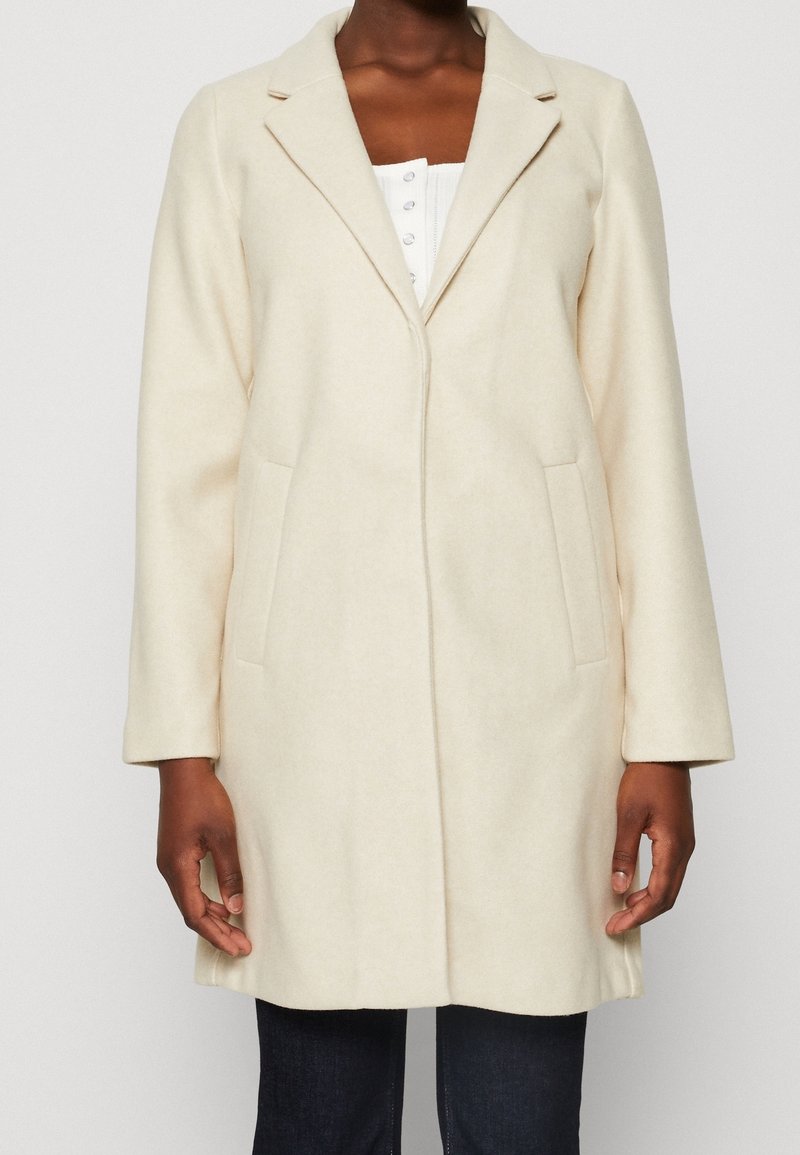 Donna che indossa un cappotto lungo color crema con revers, sopra una camicia bianca con bottoni e pantaloni scuri, in piedi su uno sfondo neutro.