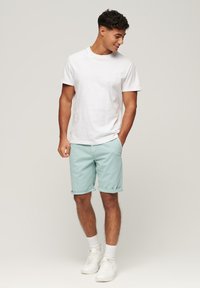 Biała bawełniana koszulka zestawiona z miętowymi szortami z podwiniętymi nogawkami. Model nosi białe trampki i skarpetki, stojąc w luźnej pozycji.