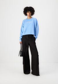 Sweatshirt blu chiaro con la scritta "Hugs", abbinato a pantaloni neri a gamba larga. Modella che tiene una piccola borsa nera, in piedi su uno sfondo semplice.