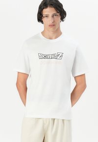 Bílý bavlněný tričko s grafikou "DRAGON BALL Z" v černé a oranžové barvě, se sedmi oranžovými kroužky pod textem, krátké rukávy.