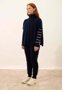 Ciemnoniebieski, oversize sweter z wysokim kołnierzem i paskowanymi rękawami; zestawiony z granatowymi legginsami i jasnobeżowymi butami sportowymi.