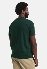 Tricou din bumbac de culoare verde închis, cu guler rotund, mâneci scurte și o textură netedă. Prezentat pe un model văzut din spate.