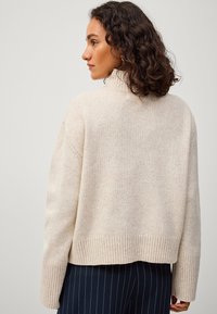 Beige gebreide trui met een hoge hals, met een geribbelde zoom en boorden. Zachte textuur, oversized pasvorm, toont de achterzijde.