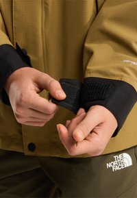 Vandtæt jakke med en tanfarvet krop og sorte ærmekanter. Har justerbar velcrolukning ved håndleddet og en diskret gittertekstur.