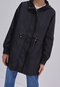 Zwarte lichtgewicht jas met capuchon, trekkoord in de taille, twee voorzakken en zilveren ritsen. Soepele textuur met elastische manchetten.