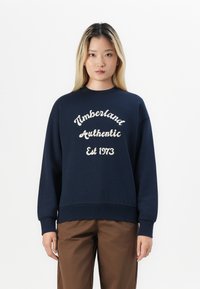 Sötétkék pulóver bordázott mandzsettával és aljjal, fehér hímzett felirattal: "Timberland Authentic Est 1973." Puha szövet textúra.