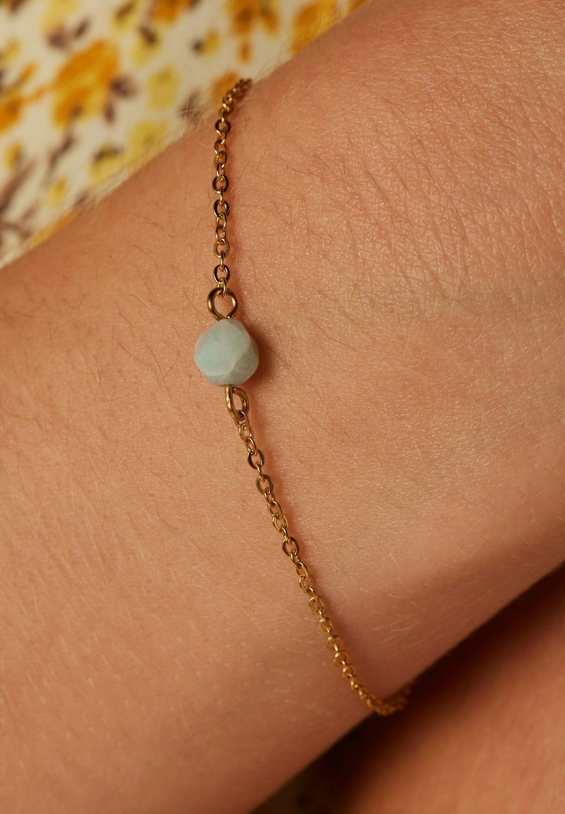 Gouden kettingarmband met een lichtblauwe onregelmatige stenen kraal, tegen een blote arm met een gestructureerd oppervlak.